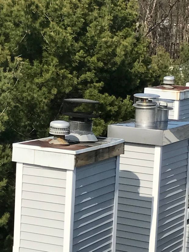Two white roof chimneys with metal caps and vents against a backdrop of trees.