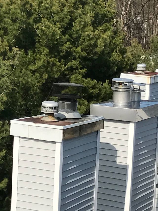 Two white roof chimneys with metal caps and vents against a backdrop of trees.