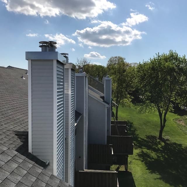 Side view of gray house chimneys and roof vents beside a green backyard tree under a blue sky
