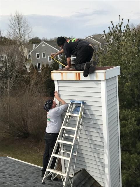 Two workers on a ladder repair the top edge of a white-sided building outdoors.