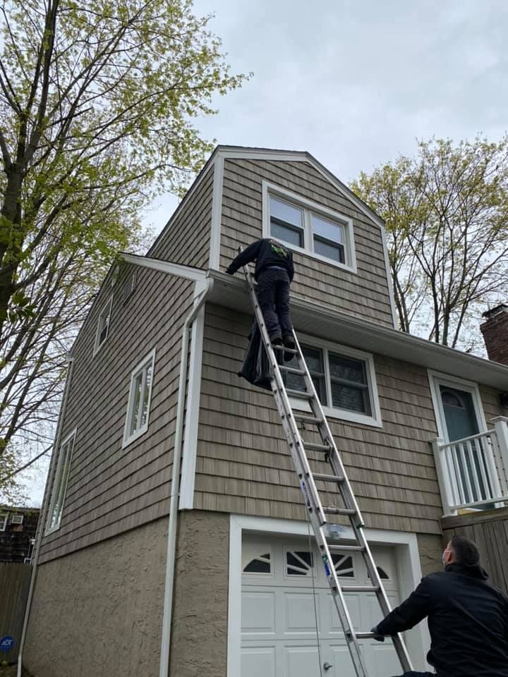Person on ladder cleaning a house roof above a garage, with another person steadying it below.