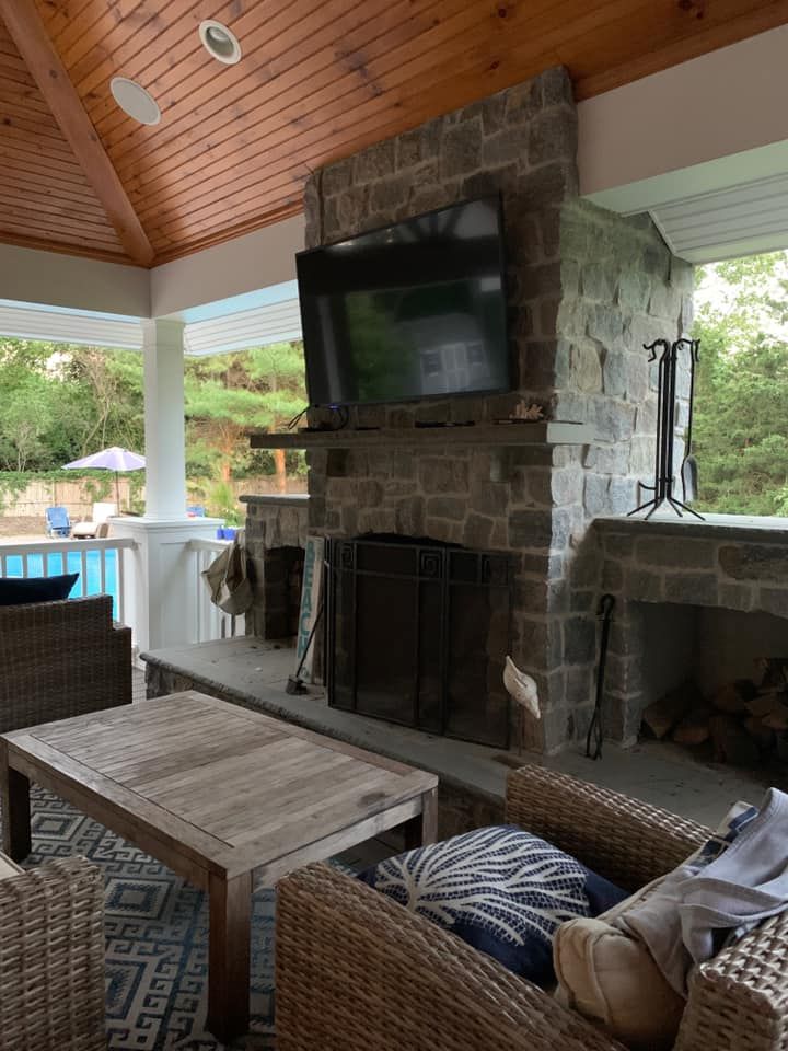 Covered patio with stone fireplace, TV, and wicker seating under a wood-paneled ceiling.