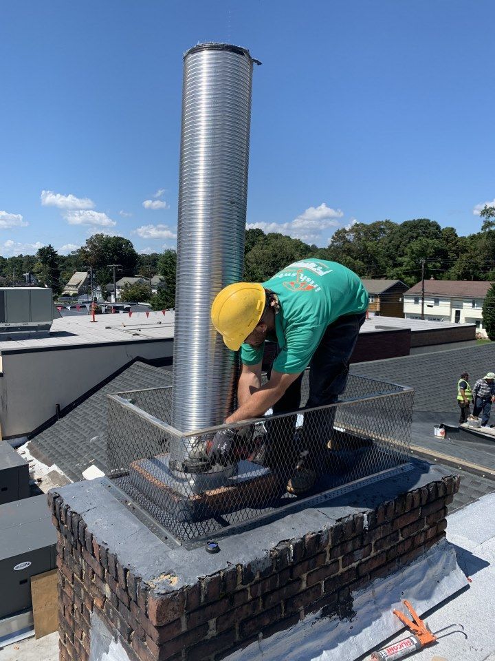 Worker in green shirt and yellow hard hat repairs a chimney on a rooftop under a blue sky