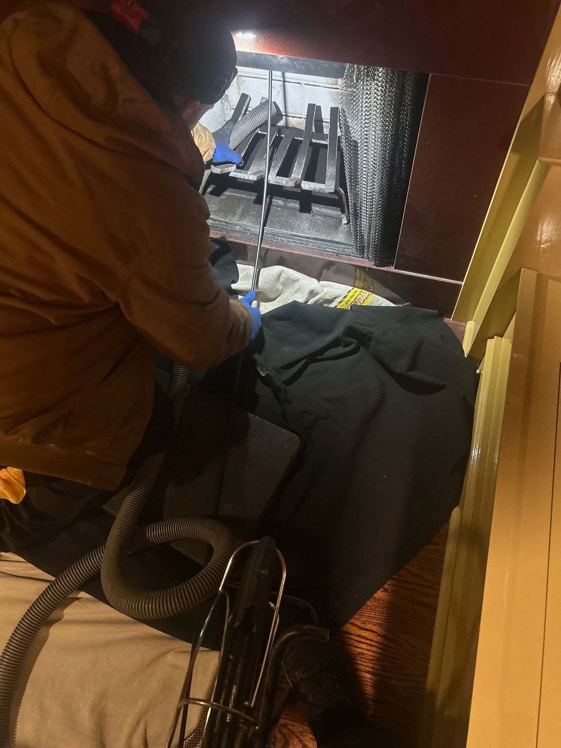 Person in an orange jacket seated indoors beside a wheelchair, looking into a metal furnace or firebox.