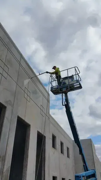 Worker on lift washing a building's exterior. Blue lift, gray wall, cloudy sky.