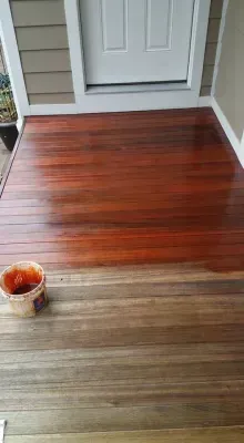 Wooden porch being stained; one half stained red-brown, the other half gray-brown. A bucket sits on the left.