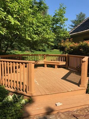 Wooden deck with built-in benches, surrounded by railing. Trees and a house are visible in the background.