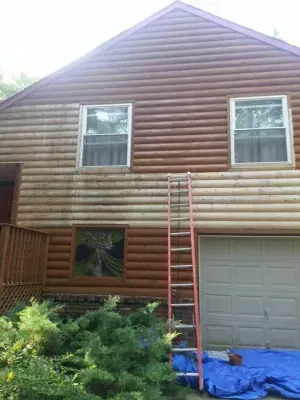 House with wooden siding; part cleaned, showing contrast. Ladder propped against wall, tarp on ground.