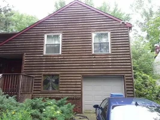 Brown log-sided house with two windows, a garage door, and a porch. A blue car is parked in front.