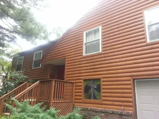Brown log-sided house with white-framed windows, a deck, and a garage door, set amidst greenery.