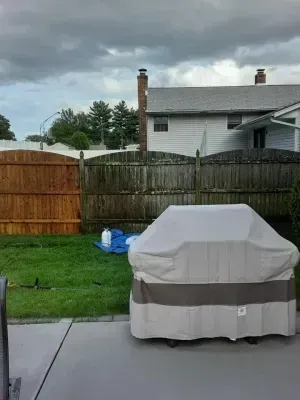 Grill covered on a patio, green lawn, wooden fences, and house under a cloudy sky.
