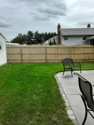 Lawn and patio in a backyard with a wooden fence, chairs, and a shed. Overcast sky.