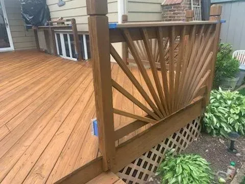 Wooden deck with decorative railing, painted brown. Backyard setting with plants and house visible.