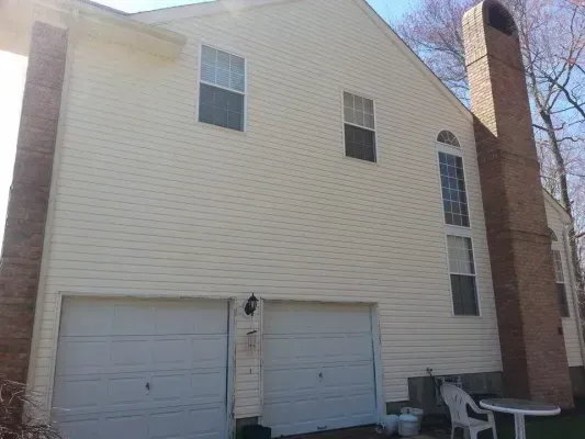 Two-car garage with beige siding, brick chimney, and windows on a sunny day.