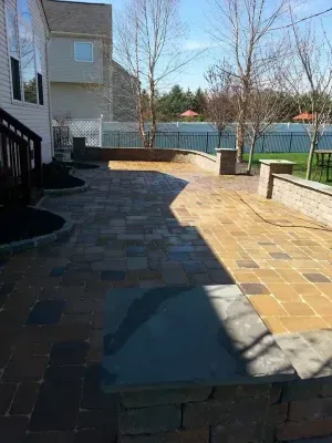 Brick patio with retaining walls, a house, and backyard with trees under a blue sky.
