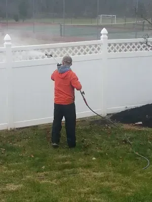 Person in orange jacket power washing a white fence outdoors near a tennis court.