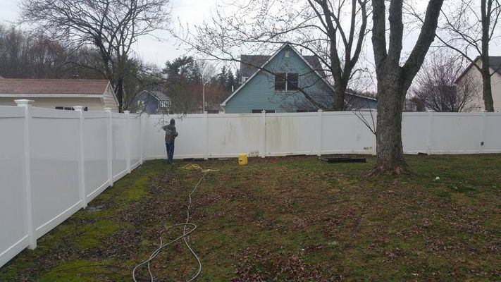 Person power washing a white vinyl fence in a backyard; some mold is visible.