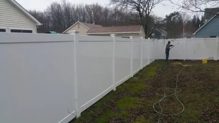 Person power washing a long white vinyl fence in a backyard.