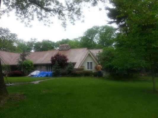 House with brown roof, surrounded by green trees and lawn, person near window.