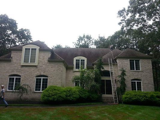 A two-story beige brick house with a dark, stained roof. A ladder leans against the building.