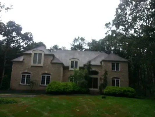 Two-story beige house with a dark roof and white-framed windows, surrounded by trees and a green lawn.