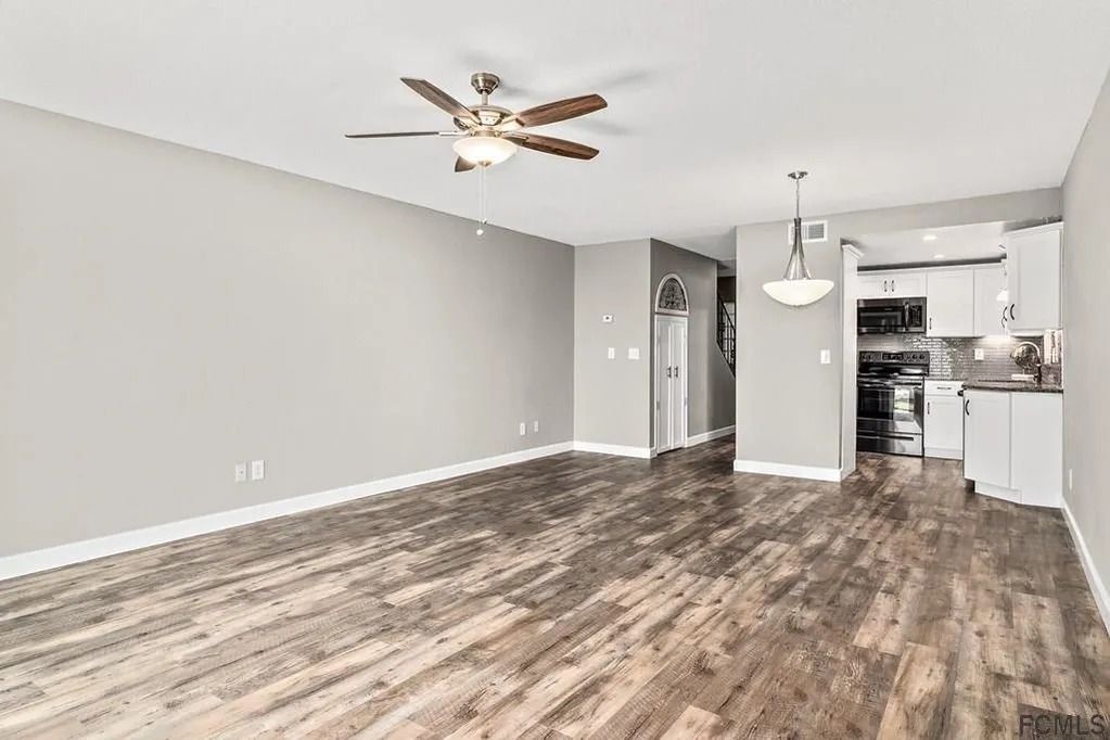 A spacious, open-concept living area with light-toned walls, wood-look flooring, a ceiling fan, and an adjacent kitchen.