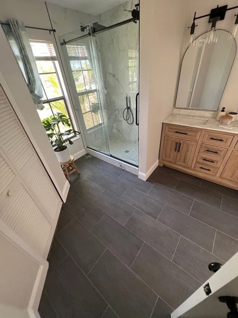 Modern bathroom with dark gray tile floors, a wooden vanity with a marble countertop, an arched mirror, and a glass shower.