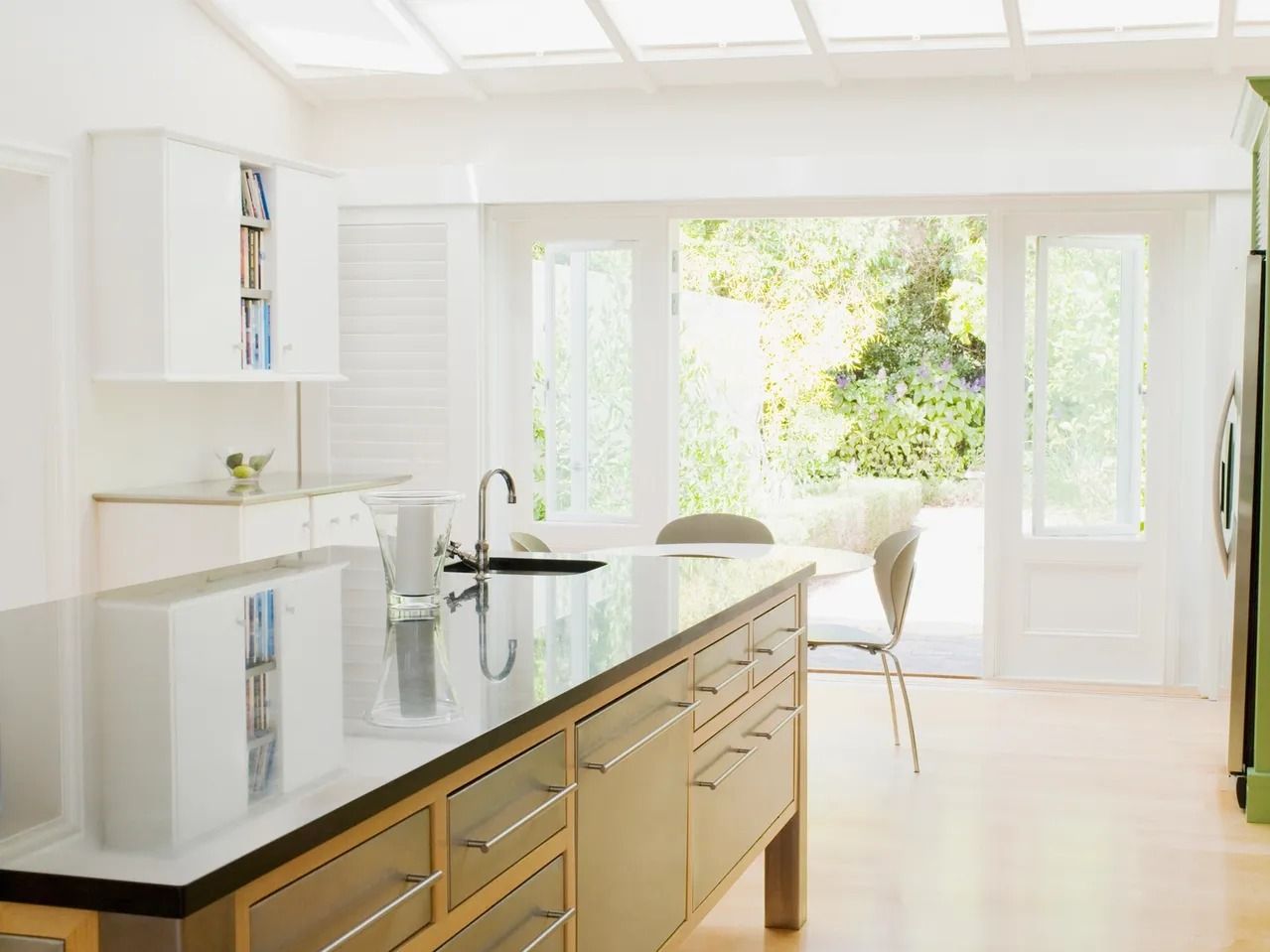 A bright, modern kitchen featuring a dark-countered island, light wood cabinets, and glass doors opening to a garden.