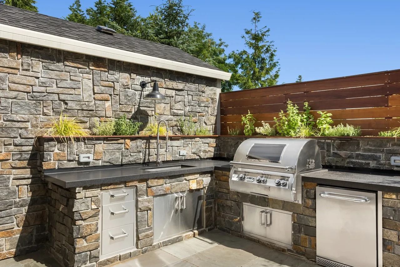 An outdoor stone kitchen area with a stainless steel grill, sink, cabinets, and a wooden privacy fence under a blue sky.