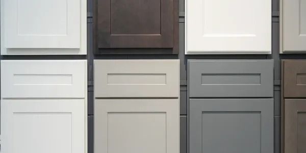 A display of Shaker-style kitchen cabinet doors and drawers in various shades of white, gray, beige, and dark brown.