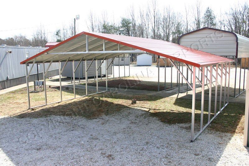 A metal carport with a red roof is sitting in a gravel lot.