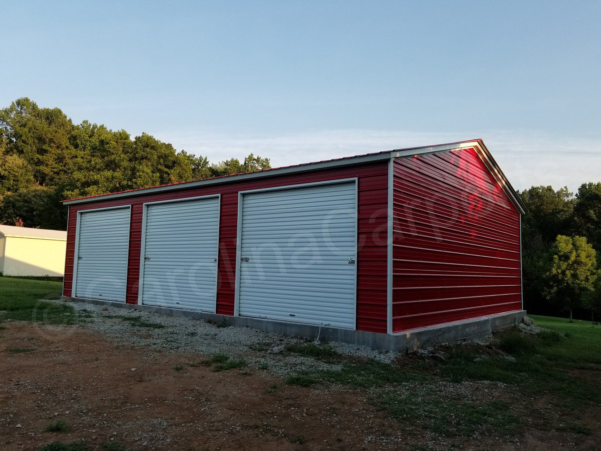 A red garage with three white doors is sitting in the middle of a grassy field.