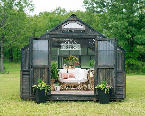 Black greenhouse with open doors, a sofa, and plants sits in a grassy field