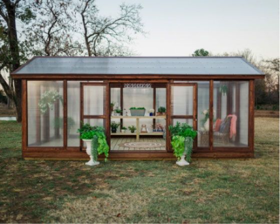 Wooden greenhouse with plants, open doors, and a cozy interior setting in a grassy yard