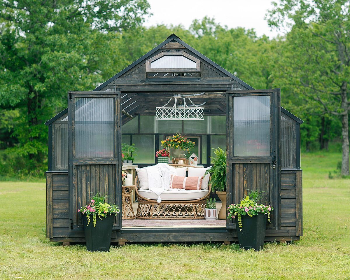 Black greenhouse with open doors, a sofa inside, and potted plants, set in a grassy field