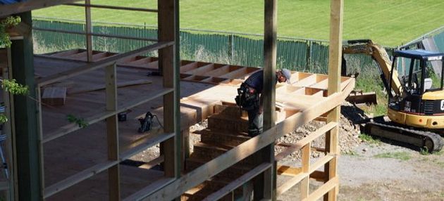 A worker framing a wooden deck structure outdoors, with a yellow excavator parked on the gravel below.