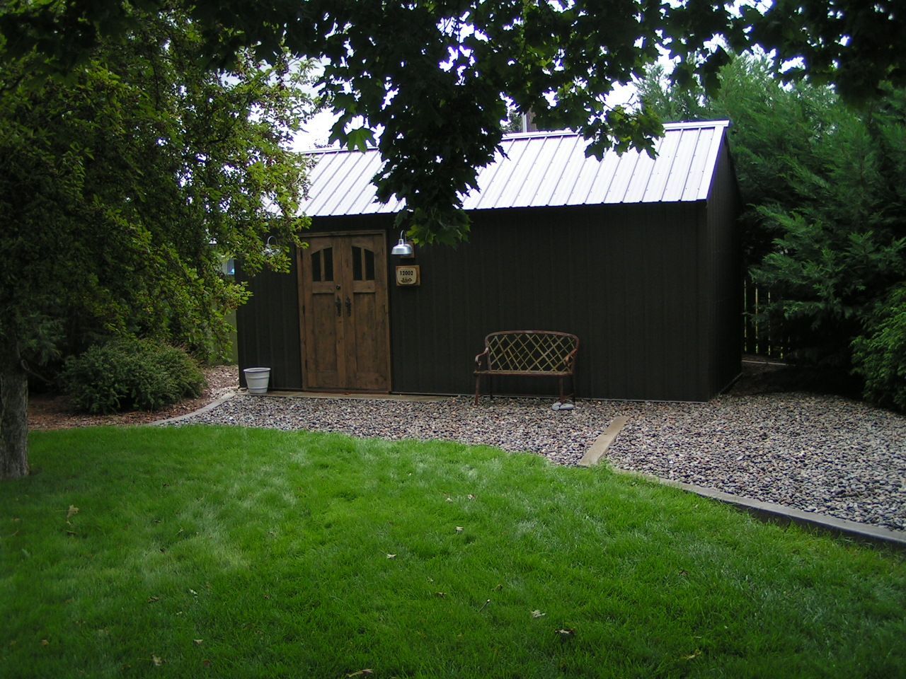 A dark brown garden shed with a silver metal roof and arched wooden double doors, set next to a gravel patio and lawn.
