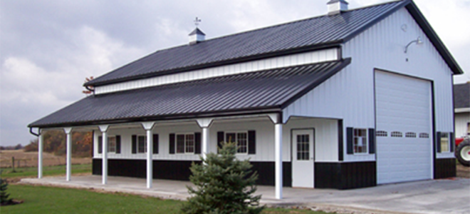A white and black pole barn building with a metal roof, a covered porch with white columns, and a large garage door.