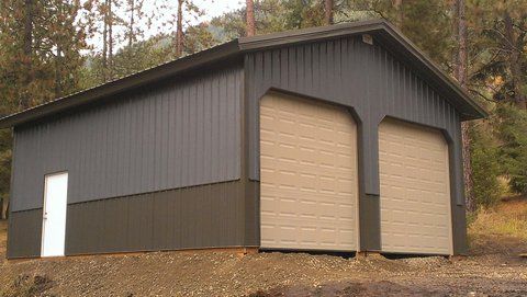 A two-car garage with dark gray vertical siding, a tan lower band, and two tan garage doors in a wooded area.