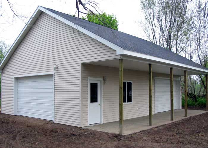 A tan, two-car garage with white trim and a covered concrete side porch set against a wooded background.