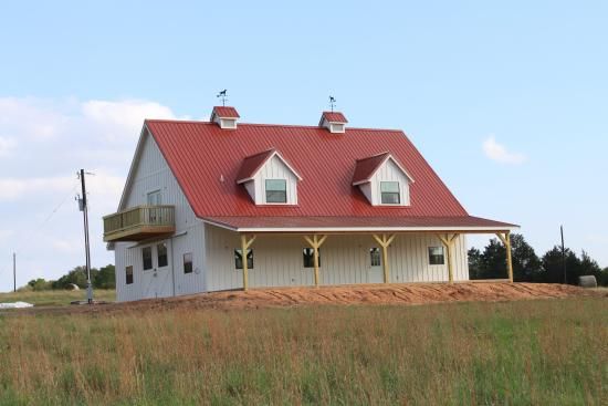 A two-story white barn-style house with a red metal roof, dormer windows, and a front porch, set in an open field.