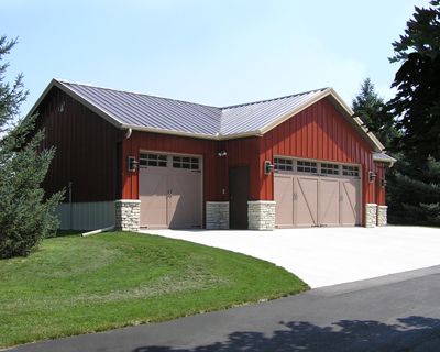 A red L-shaped garage with a metal roof, stone accents, and tan carriage-style doors on a paved driveway near a lawn.