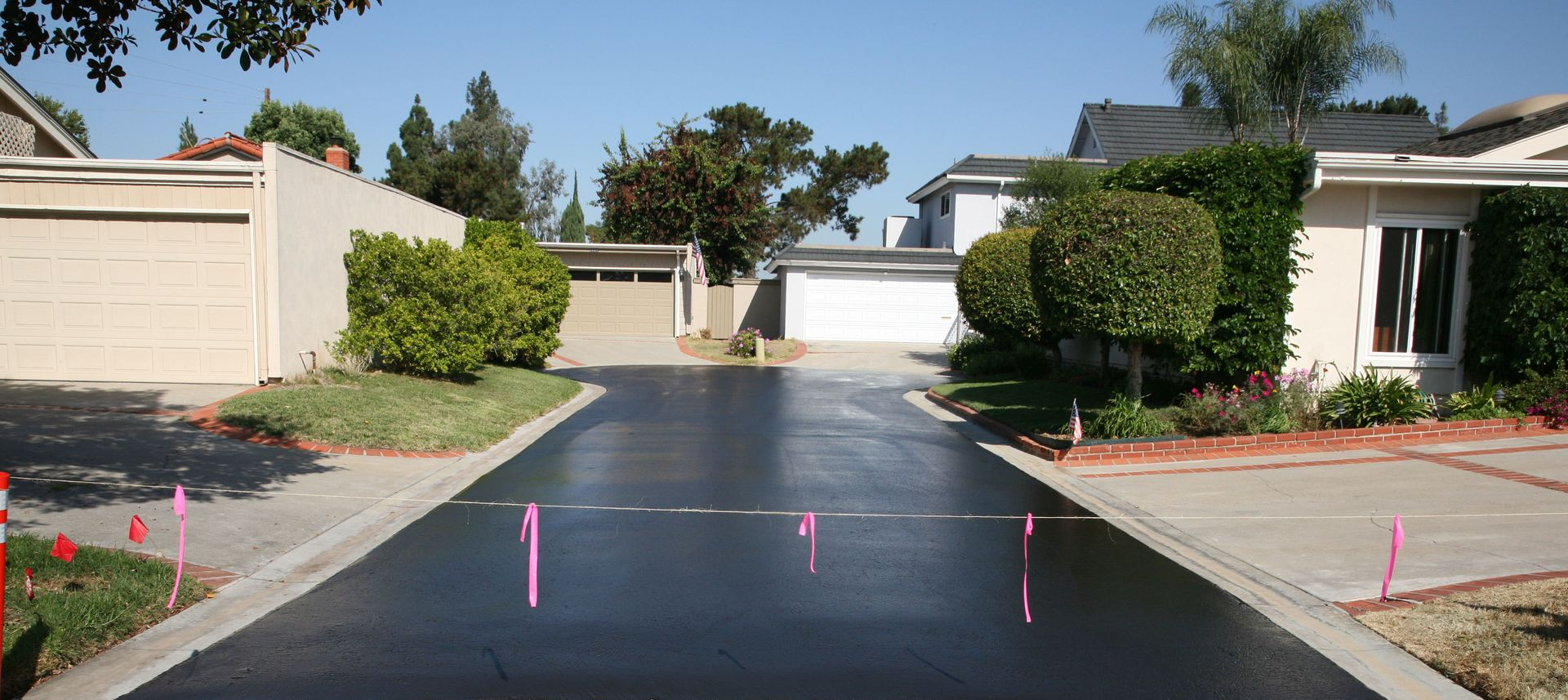 Driveway with recently paved black asphalt. Homes and trees line the sides. Pink markers dot the drive.