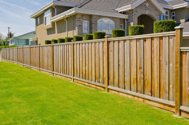 Wooden fence bordering a well-manicured green lawn next to a two-story beige house.