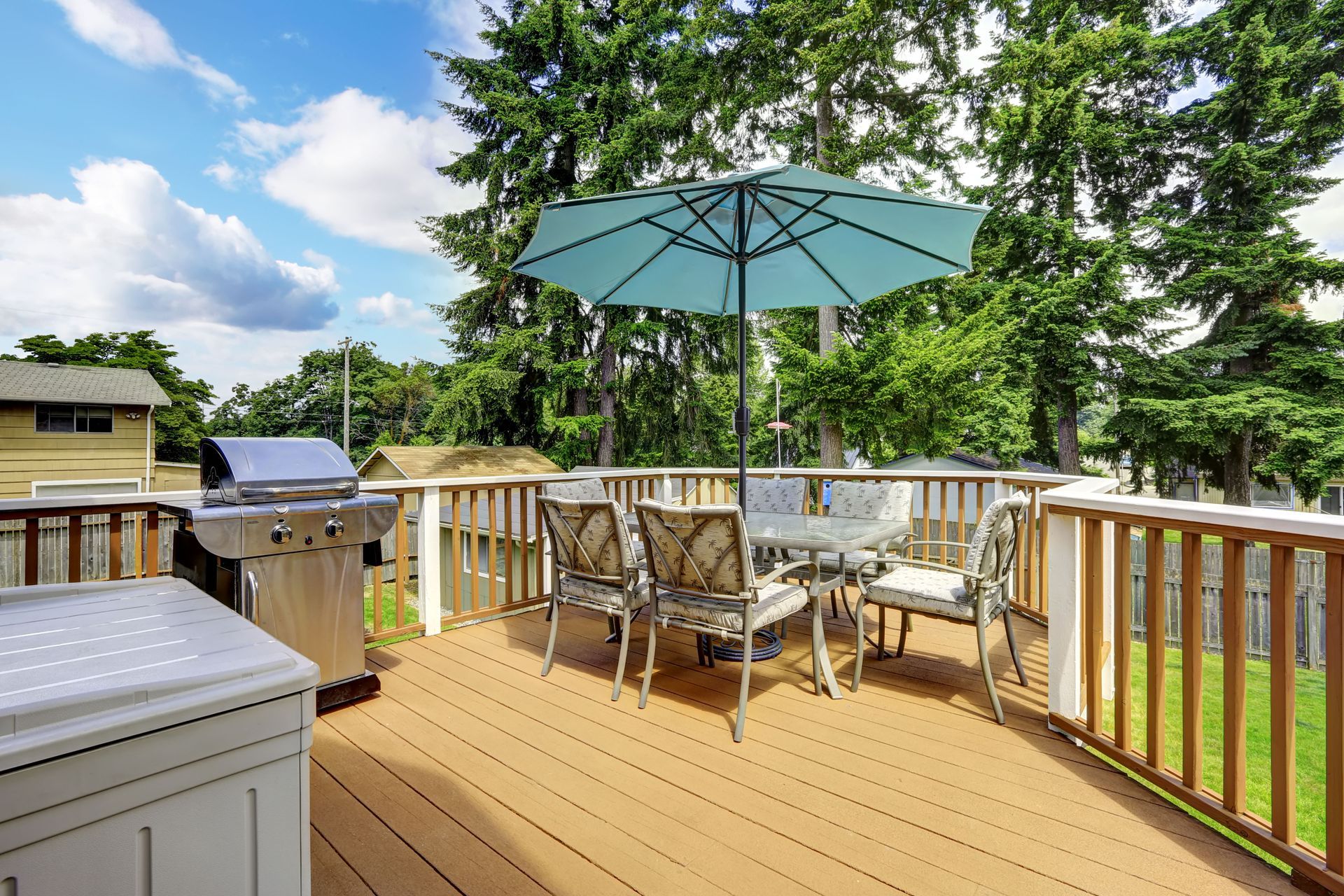 Wooden deck with outdoor dining set and grill under a teal umbrella; green trees and blue sky background.