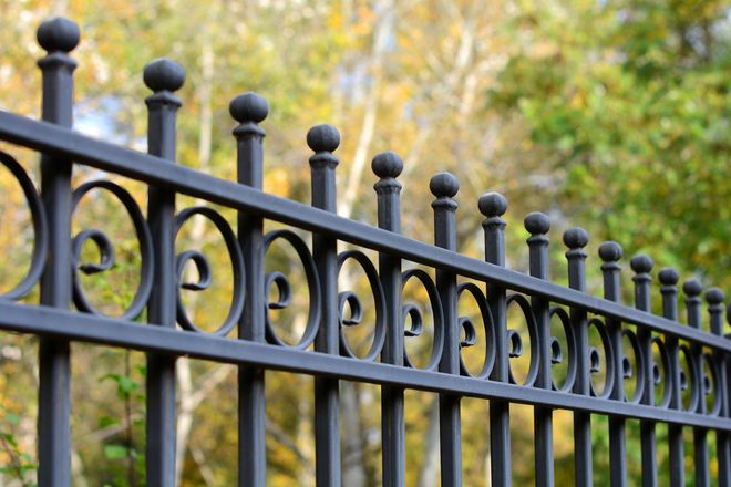 Black wrought-iron fence with decorative swirls and ball finials, blurred background of trees.
