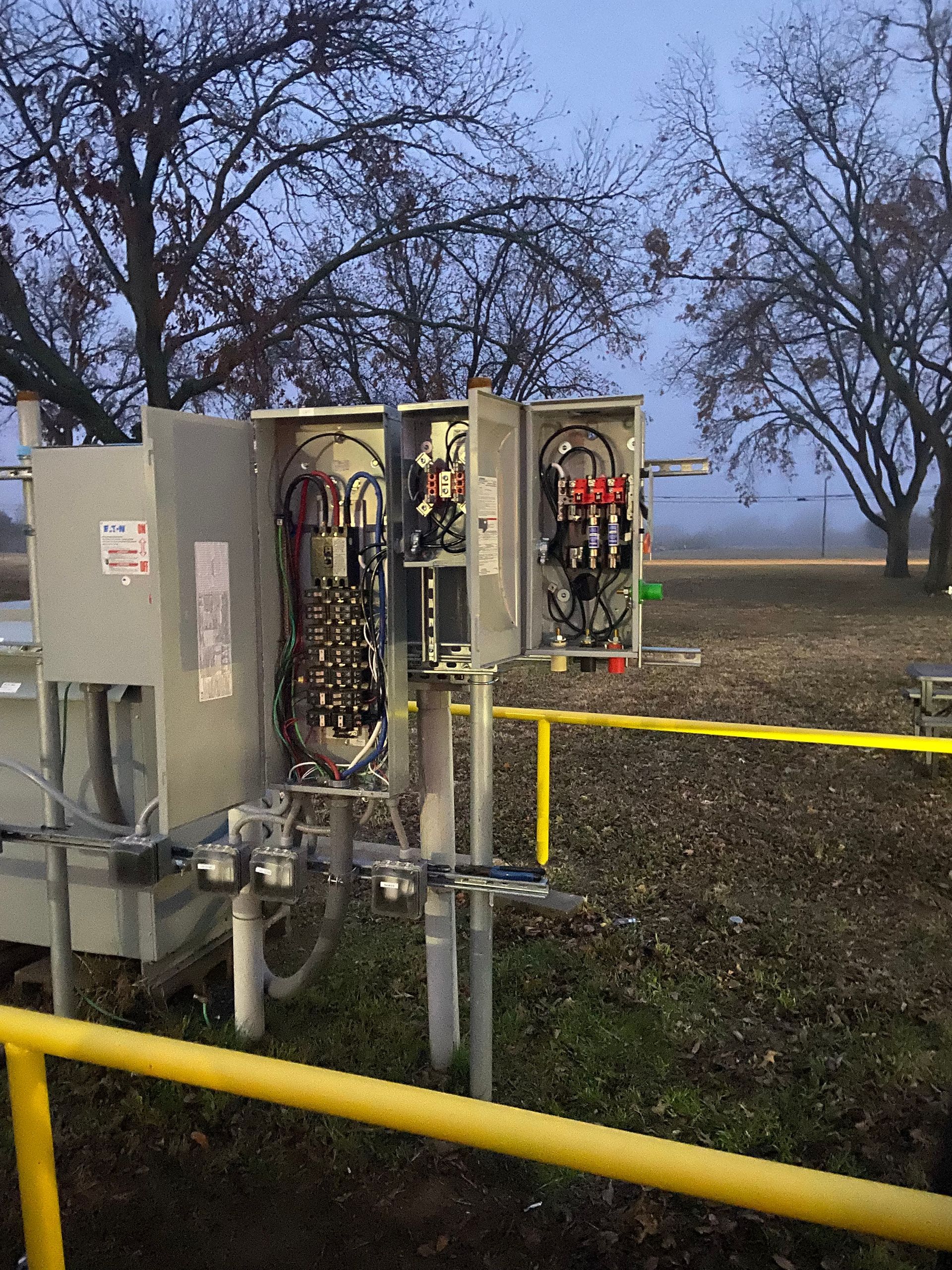 Two electrical boxes are sitting in a field next to a yellow railing