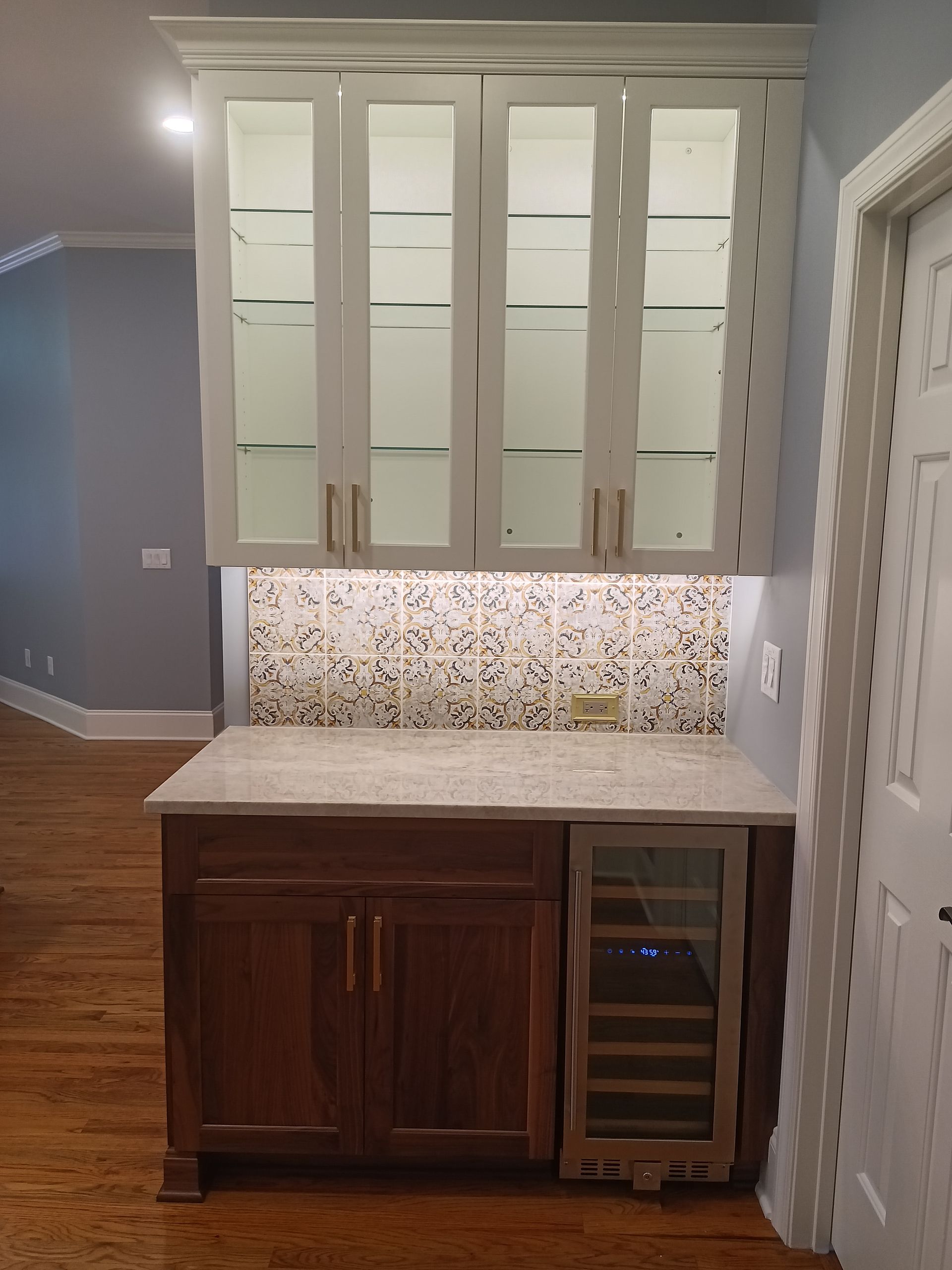 A small home bar with upper glass-door cabinets and a lower wood cabinet, includes a wine fridge, and stone countertop.