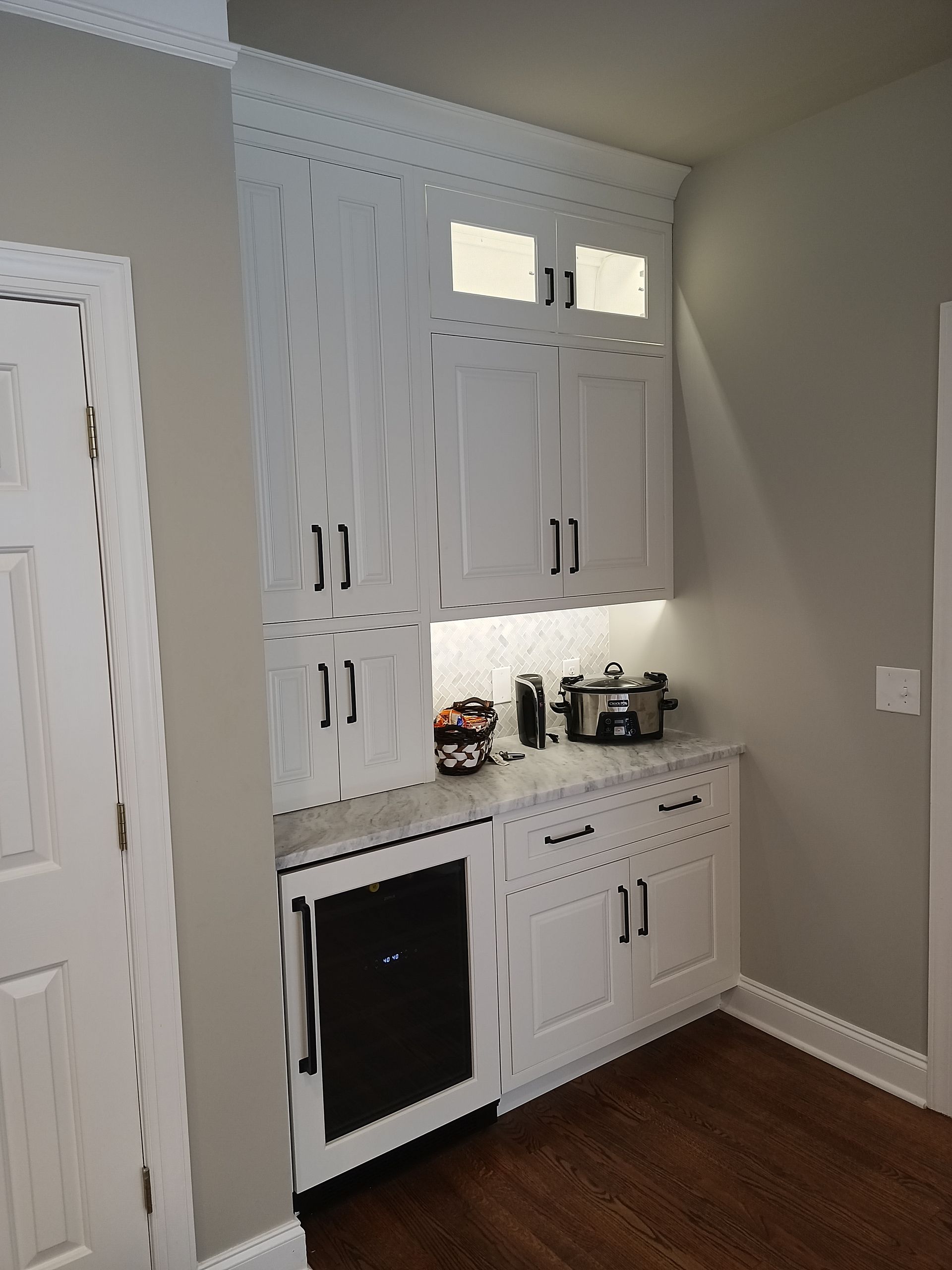 White built-in coffee bar with cabinets, countertop, and appliances, next to a door, on hardwood floor.