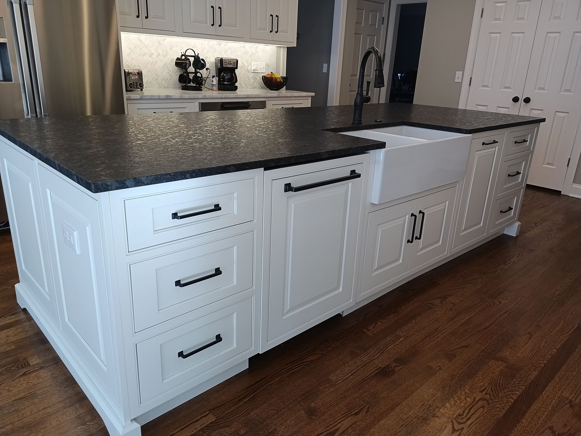 White kitchen island with black countertop, farmhouse sink, and drawers; hardwood floor.